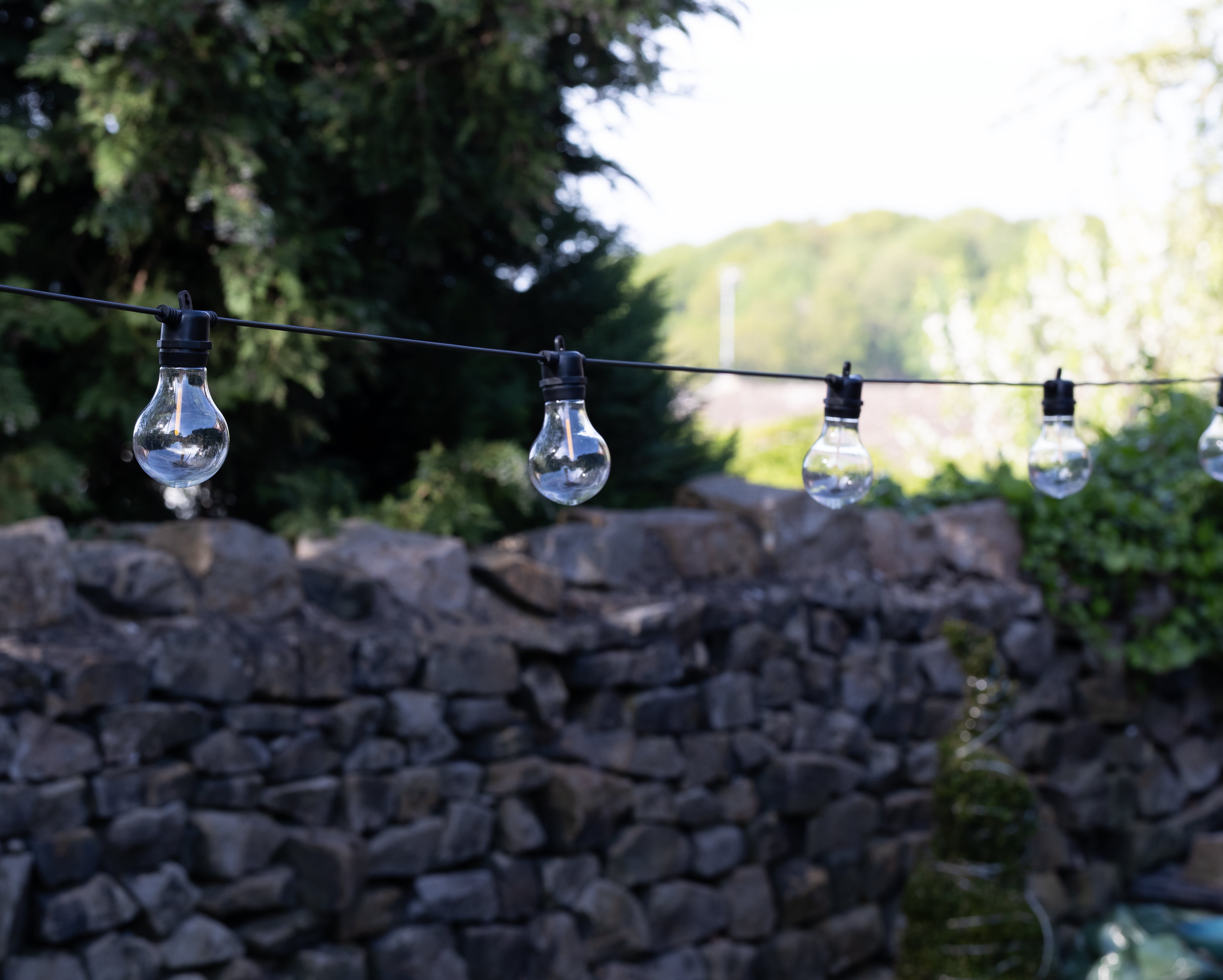 String of light bulbs hanging above a stone wall with trees in the background