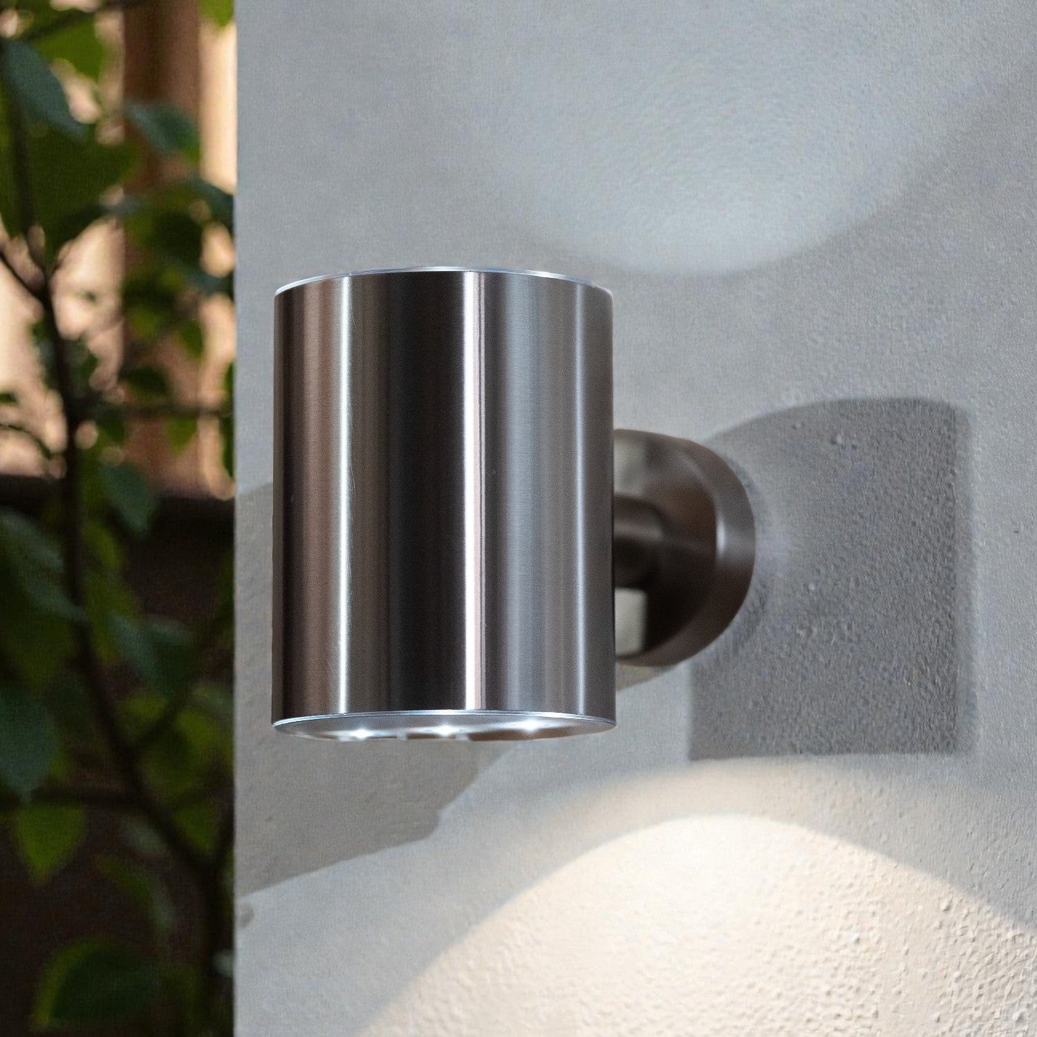 Metallic outdoor wall light fixture on a textured wall with plants in the background