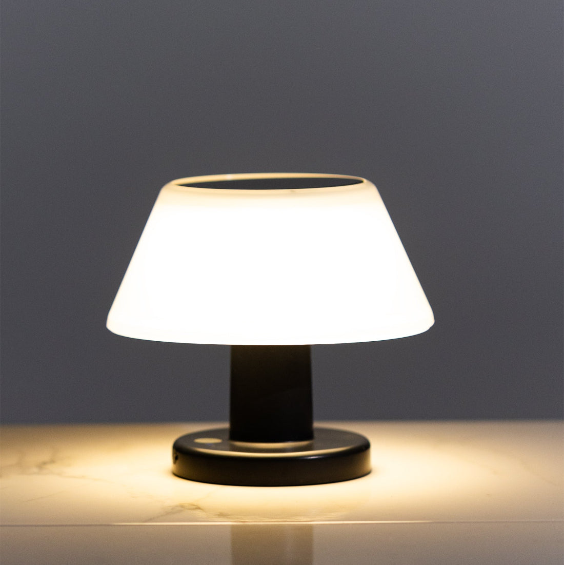 Outdoor solar table lamp with warm white LED, placed on a table next to a glass vase with flowers, with a dark background.