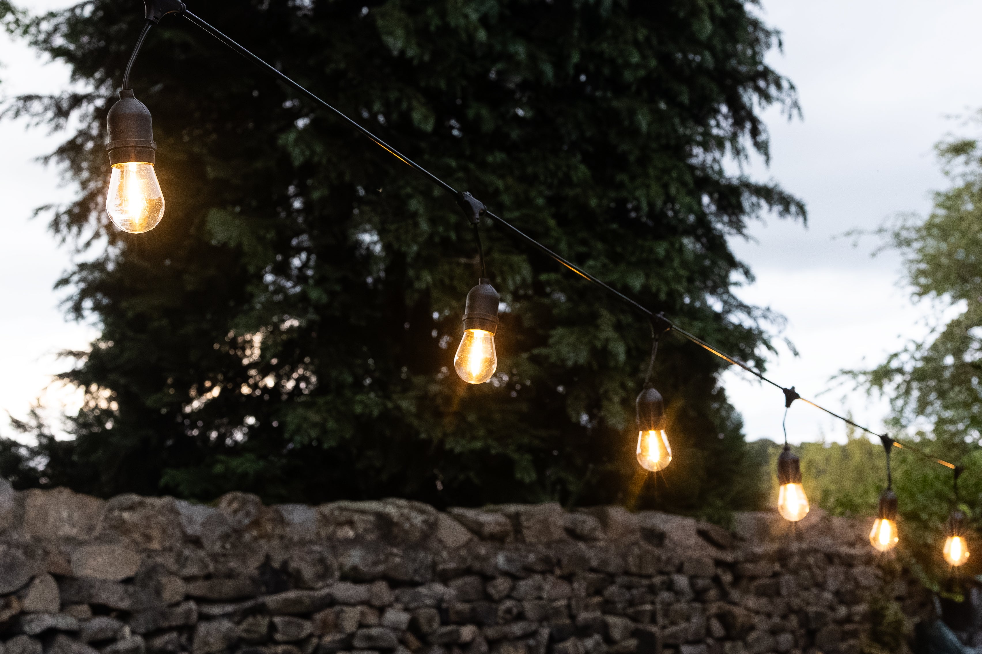 String of light bulbs hanging above a stone wall with trees in the background
