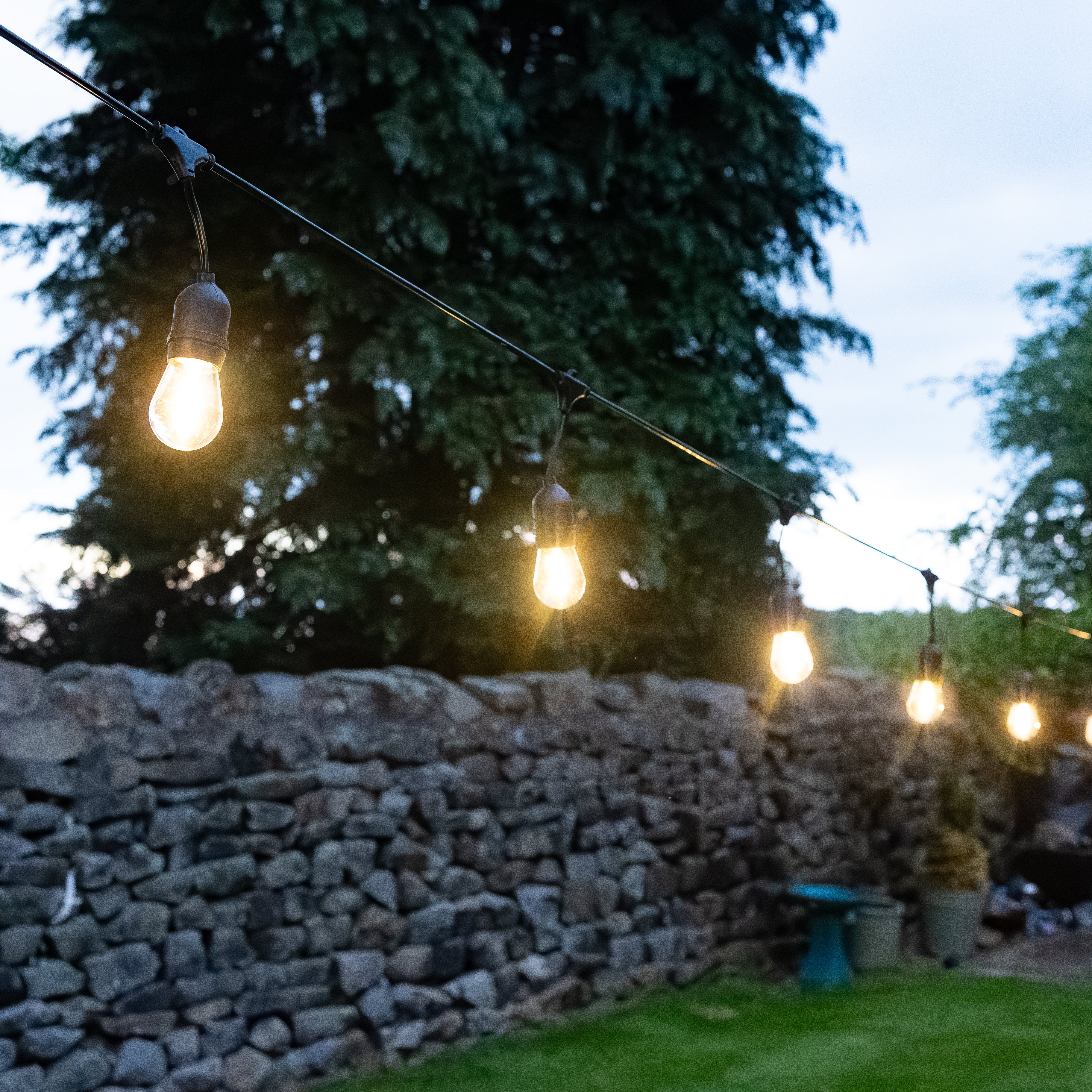 String lights hanging above a stone wall with trees in the background