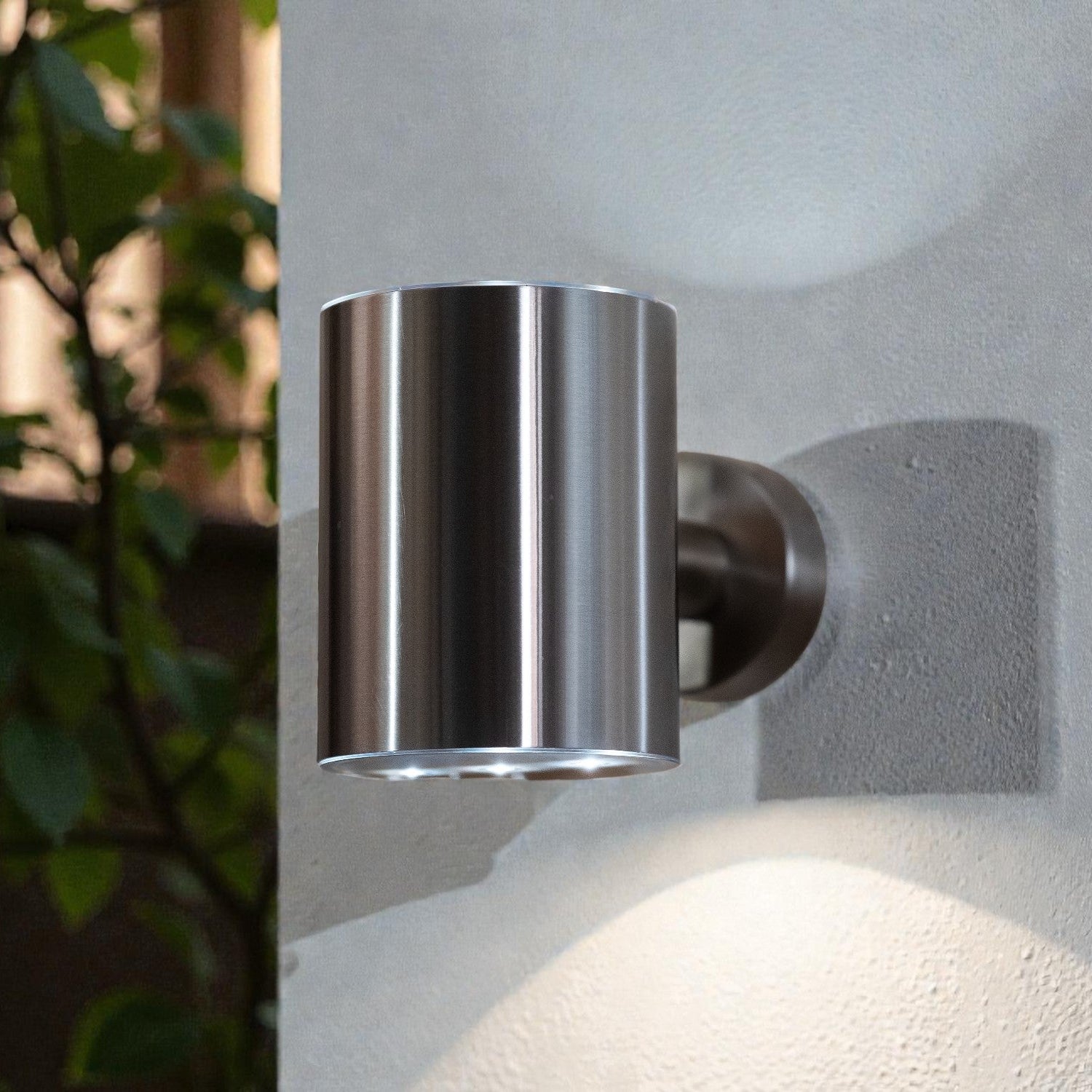 Metallic outdoor wall light fixture on a textured wall with plants in the background